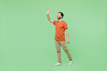 Full body side view smiling happy young man 20s wearing casual orange t-shirt walking going waving hand isolated on plain pastel light green color background studio portrait. People lifestyle concept.