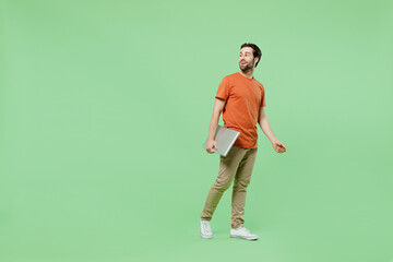 Fototapeta na wymiar Full body young fun man 20s wear orange t-shirt hold closed laptop pc computer look aside on workspace area copy space isolated on plain pastel light green background studio. People lifestyle concept.