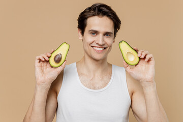 Attractive happy fun young man 20s perfect skin wear undershirt hold two halfs of avocado isolated...