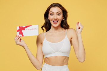 Happy european young brunette woman 20s in white underwear with perfect fit body hold gift certificate coupon voucher card for store do winner gesture isolated on plain yellow color background studio