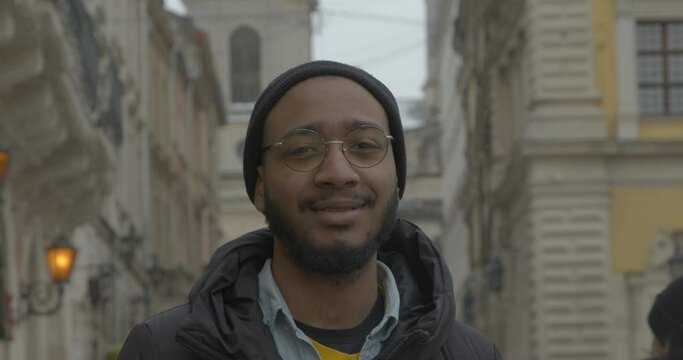 African American man in glasses and hat smiles standing in street. Young tourist walks against vintage buildings in old city on winter day hero shot slow motion