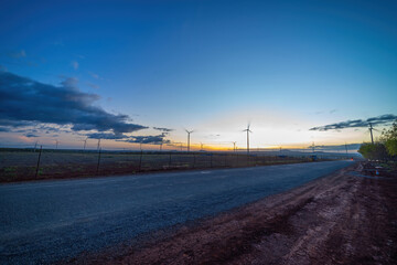 Fototapeta premium Wind turbine farm and agricultural fields on a summer day. Wind electricity generator in sunset sky, power plant, wind efficiency.