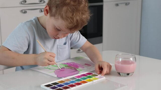 Cute boy enjoys drawing with a brush and pink paint while sitting at a white table at home in kitchen. Child learns to draw with watercolor paints. First steps of a Caucasian boy in the visual arts.