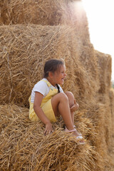 Naklejka na ściany i meble Super happy young girl laughing out loud smiling with teeth with closed eyes sitting on haystack wearing sundress. Having fun away from city on field full of golden hay.
