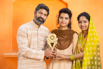 Proud traditional indian parents with young daughter holding winning prize celebrating victory. Happy young school girl showing trophy standing with her father and mother.