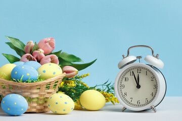 Easter concept. Basket of eggs alarm clock flowers on a blue background copy space.