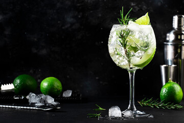 Gin tonic alcoholic cocktail drink with dry gin, rosemary, tonic, lime and ice cubes in wine glass. Black bar counter background, bar tools, copy space - obrazy, fototapety, plakaty