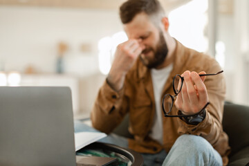 Tired Male Holding Glasses Massaging Nosebridge Sitting At Laptop Indoors