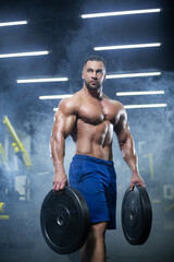 Fototapeta na wymiar Handsome muscular athlete man is standing in a gym against the background of smoke holding barbell discs in both hands