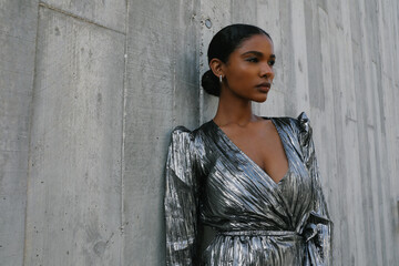 Young African American woman posing on the grey wall wears silver dress.
