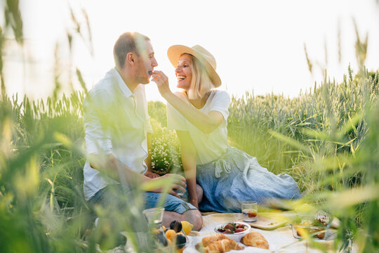 Happy young family enjoy and laughing on picnic in green wheat field at nature. Adult blond woman feeding her husband with a cherry berry. Romantic date surprise concept.