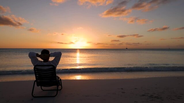 A happy vacation man is feeling free and relaxes in a sun chair on a tropical beach during colorful sunset time