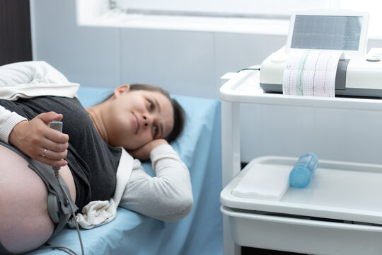Pregnant Woman On Hospital Couch During Medical Control Cardiotocography. Gynecologist Doctor Hands Turn On Adjust CTG Device Clinic Consultant. Examine Expectant Belly Baby Mother Healthcare Check Up