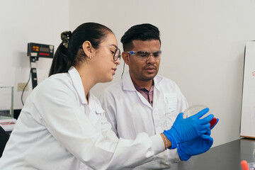 Laboratory technicians examining growths on cell culture plates. They are seen taking safety precautions by wearing goggles, gloves and lab coats.
