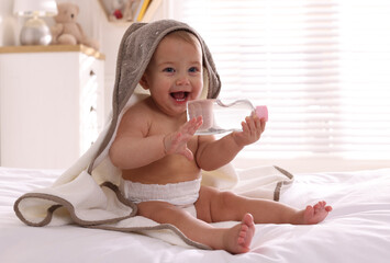 Cute little baby with towel and bottle of massage oil on bed at home