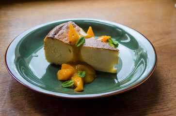 Piece of cheesecake on a green plate with peach slices and mint leaves and sea buckthorn glaze on a wooden background.