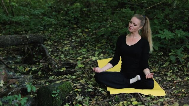 Beautiful middle aged woman practice yoga in autumn forest near river. Female seats on yellow yoga mat on ground in covered by dry leaves woodland and doing exercises.