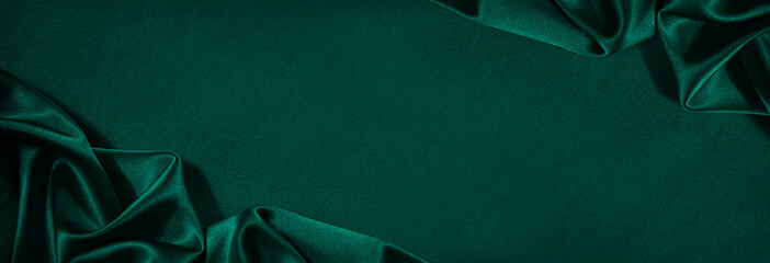Dark green silk satin background. Beautiful soft folds on the smooth surface of the fabric. Luxury background with copy space for design. Wide banner. Top view. Flat lay.