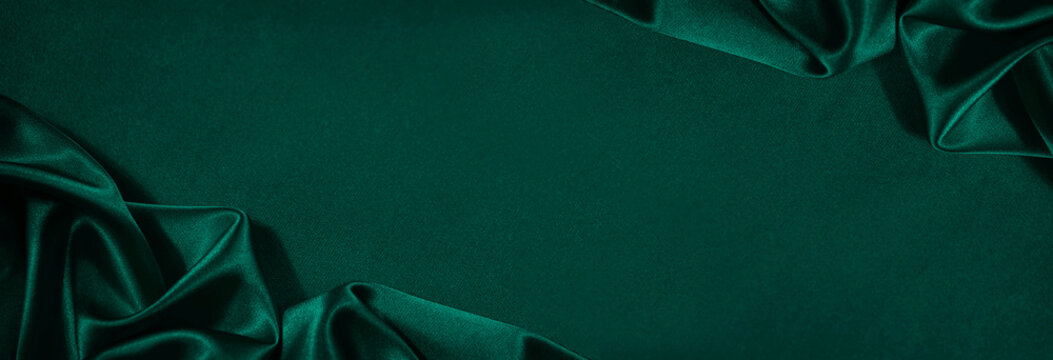 Dark green silk satin background. Beautiful soft folds on the smooth surface of the fabric. Luxury background with copy space for design. Wide banner. Top view. Flat lay.