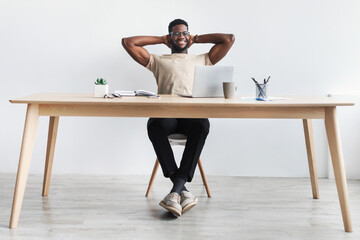 Smiling black company employee leaning back on chair with hands behind head, relaxing after online...