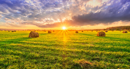 Scenic view at picturesque burning sunset in a green shiny field with hay stacks, bright cloudy sky , golden sun rays and road leading far away, summer valley landscape