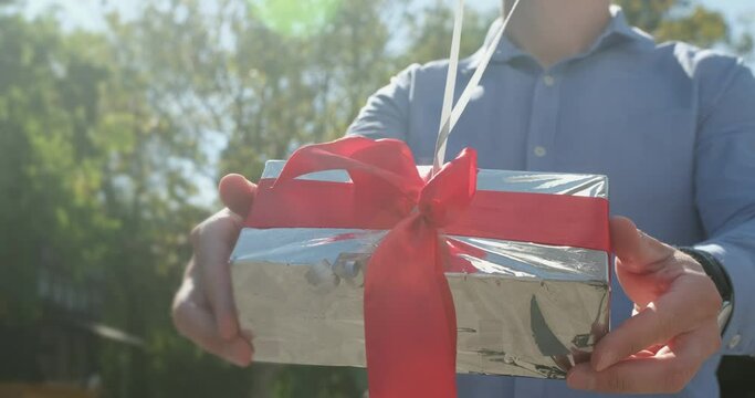 Man shows silver present box with red ribbon attached to helium balloons on sunny day. Inflatable balls ready to fly up in air close view