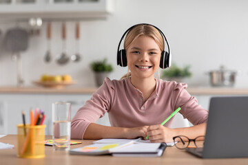 Cheerful european teen girl blonde in headphones study at home at table with laptop in kitchen interior