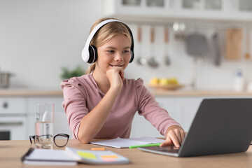 Glad happy caucasian young girl blonde in headphones at table with laptop in kitchen interior, free space