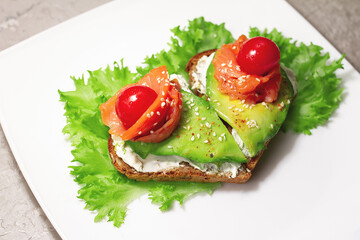 healthy snack of heart shaped grain bread curd cheese, avocado and salmon with lettuce and cherry tomatoes, sesame on white plate