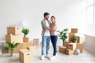 Glad european millennial wife and husband in casual hugging in room with cardboard boxes, enjoy buying of new apartment