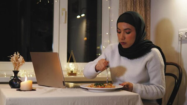 Muslim arabic young woman in hijab staying at home, having dinner, working on laptop at night. Tired businesswoman freelancer watching on laptop screen 
