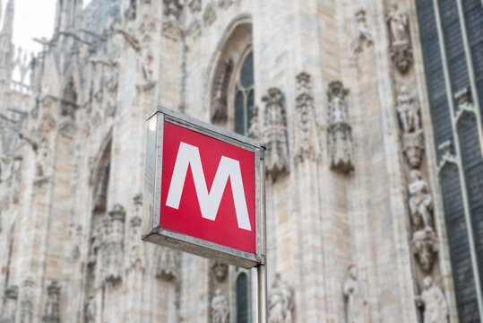 Red metro sign at subway station, M letter. Urban transport concept. Piazza del Duomo, with the blurred Milan Cathedral in the background, Italy