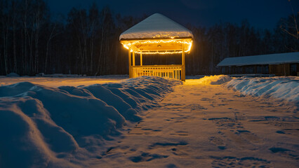 Gazebo, éclairé par des lanternes, dans le parc d& 39 hiver. Neige sur le toit, traces de pas sur le chemin. Il y a des congères sur les bords des routes. Au loin, dans l& 39 obscurité, un bosquet de bouleaux est visible. Sibérie