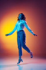 Fototapeta premium Full-length portrait of happy young beautiful girl with long curly hair jumping isolated on orange background in neon light, filter. Concept of emotions, beauty, fashion