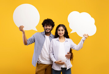 Portrait of happy indian pregnant couple holding empty speech bubbles, guy hugging wife, standing...