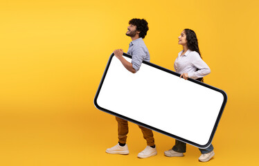 Young indian couple holding giant smartphone with mockup for design, yellow studio background, free...