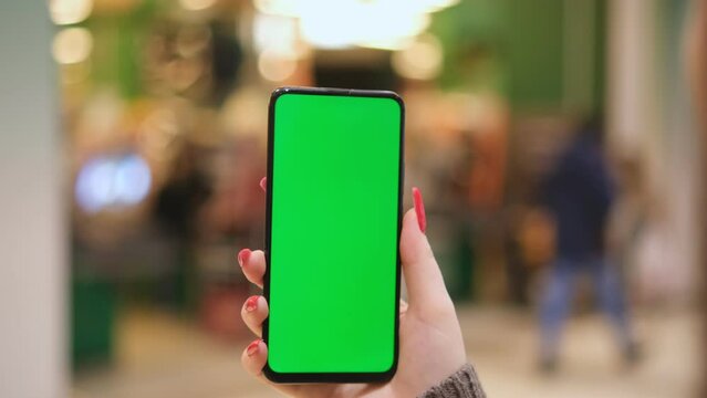 young woman holding a phone with chroma key on the background of a supermarket. The girl is holding a smartphone with a green screen and in the mall. Business woman working on smartphone surfing the