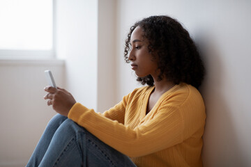 Depressed young black woman sitting near wall, staring at smartphone, suffering from apathy at home