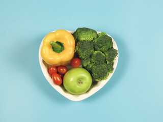 flat lay of vegetables  capsicum, broccoli tomatoes and green apple in heart shape plate  on blue background with copy space, healthy lifestyle and weight loss concept.
