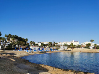 A small sparsely populated sandy beach with sun loungers and umbrellas in the bay of the Mediterranean Sea on a cloudless day.