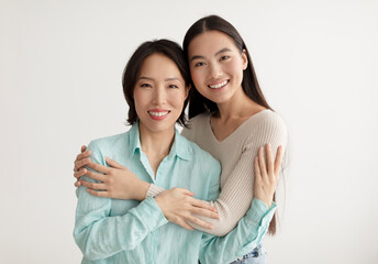 Joyful young Asian woman and her senior mother hugging and smiling at camera on white studio...