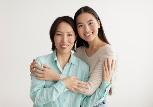 Joyful young Asian woman and her senior mother hugging and smiling at camera on white studio background