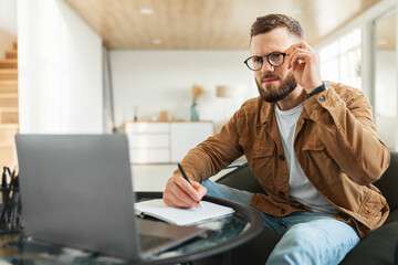 Businessman Looking At Laptop Touching Eyeglasses Taking Notes At Home