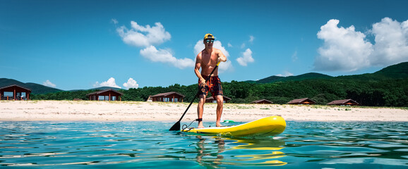 Tanned man stands on a yellow paddle board and paddles. Active sport on the beach in the turquoise...