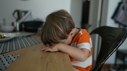 Shy Child covering face with hand little boy sitting at table feeling bore