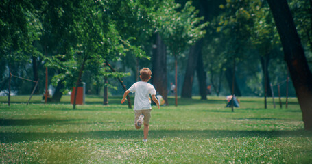 Energetic elementary age boy run at green lush field on sunny day. Camera zoom.