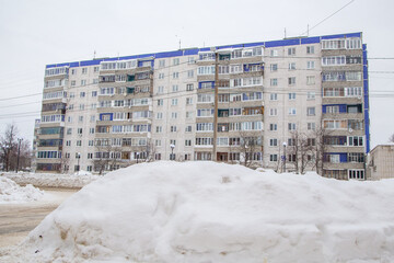 A huge big snowdrift by the road against the backdrop of city houses. On the road lies dirty snow in high heaps. Urban winter landscape. Cloudy winter day, soft light.