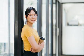 Happy young female in trendy outfit holding cup of coffee and using smartphone while standing at table of coffee shop