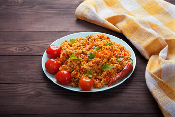 Jollof rice, tomatoes and hot peppers on a blue plate, linen napkin on a brown wooden background. National cuisine of Africa.