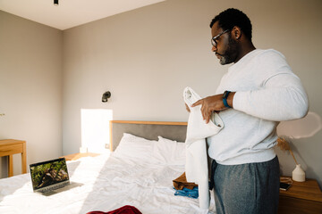 Black man using laptop while folding his clothes in bedroom